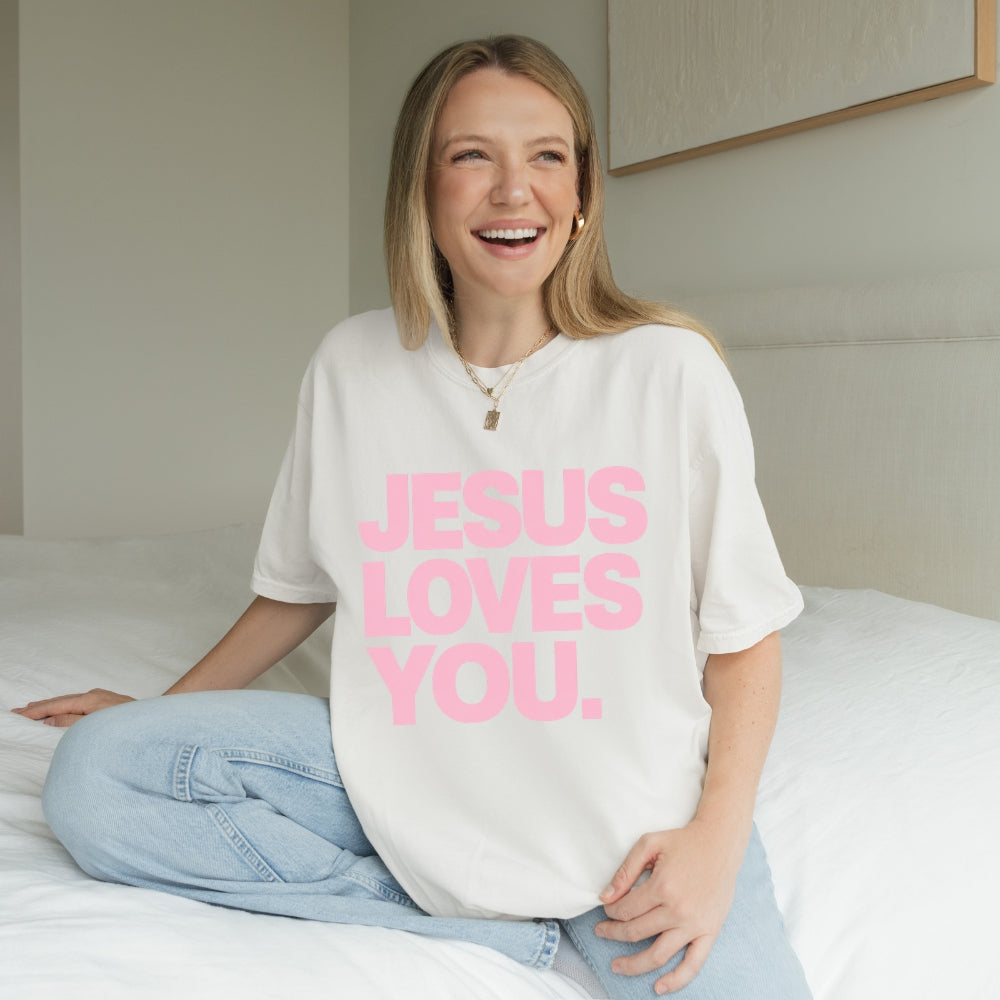 A smiling woman sitting on a bed wearing a white oversized Everyday Faith T-shirt with the phrase 'JESUS LOVES YOU.' printed in large pink letters, paired with light blue jeans.