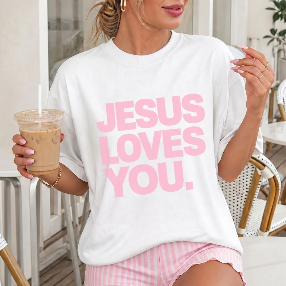 A woman wearing a white oversized Everyday Faith graphic T-shirt with the phrase 'JESUS LOVES YOU.' printed in large pink letters, holding an iced coffee in a bright indoor setting.