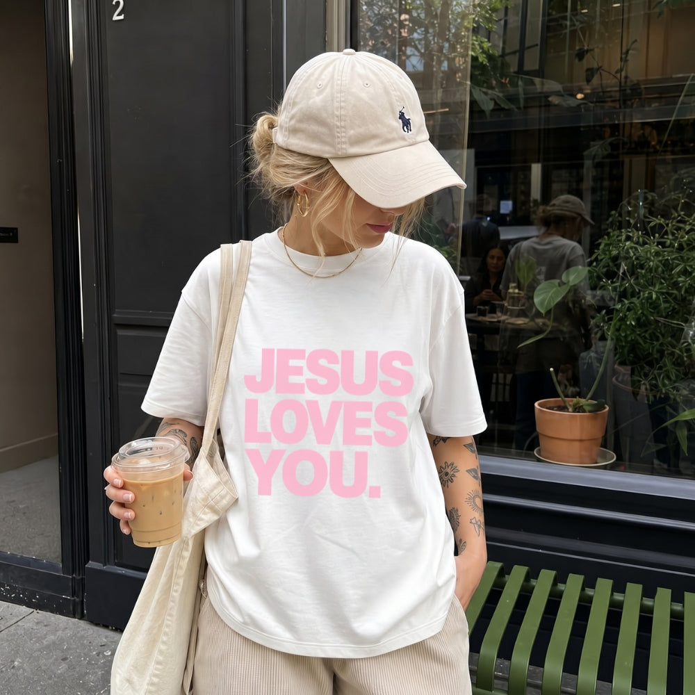 A woman wearing a beige baseball cap and a white Everyday Faith graphic t-shirt with the text 'JESUS LOVES YOU.' in pink, standing on a city street next to a cafe window and holding an iced coffee.