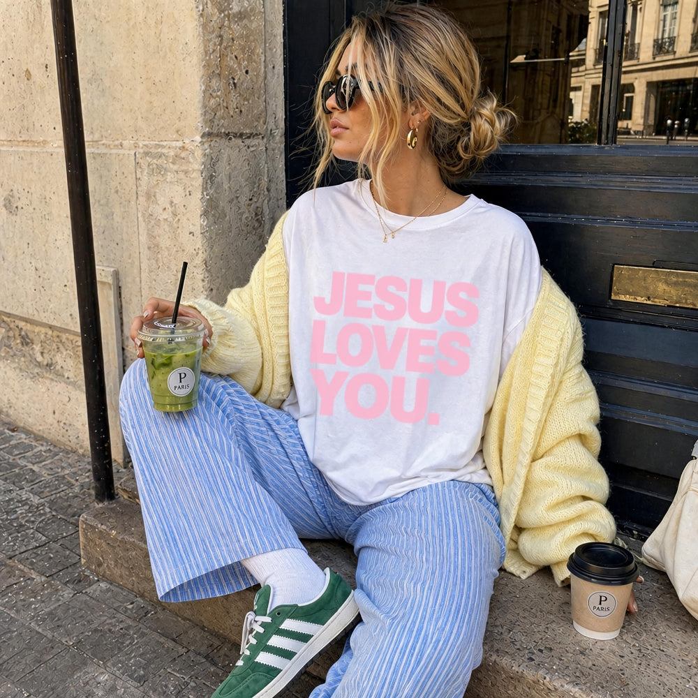 A woman sitting on stone steps outdoors wearing a white Everyday Faith graphic T-shirt with pink text reading 'JESUS LOVES YOU.' She has on a pale yellow knit cardigan, blue-and-white striped pants, green sneakers, and holds an iced drink and a coffee cup.