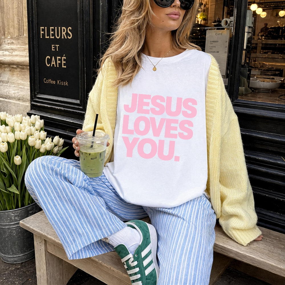 A woman sits on a wooden bench outside a cafe wearing a white Everyday Faith graphic t-shirt with pink text that reads 'JESUS LOVES YOU.' She is styled with a pale yellow cardigan, blue and white striped pants, green sneakers, and holds an iced matcha drink.