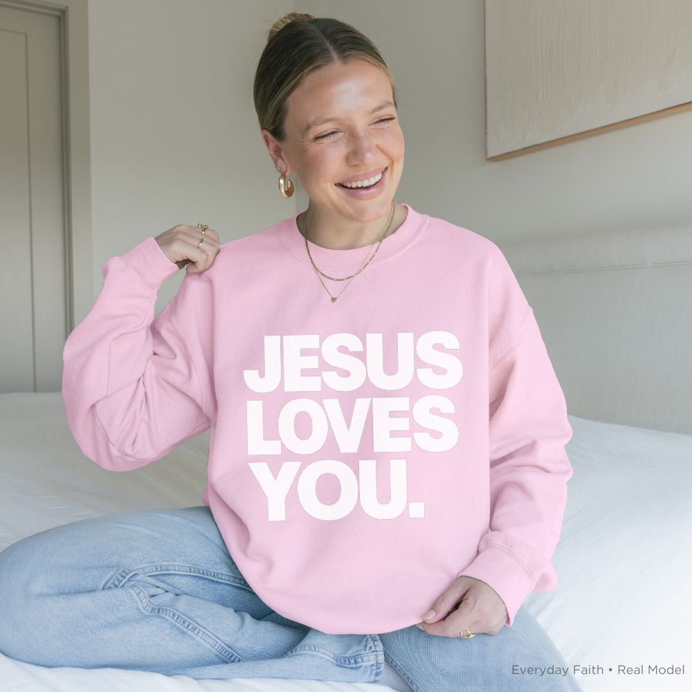 A person sitting on a bed wearing a light pink Everyday Faith sweatshirt with the text 'JESUS LOVES YOU.' printed in bold white capital letters across the front.