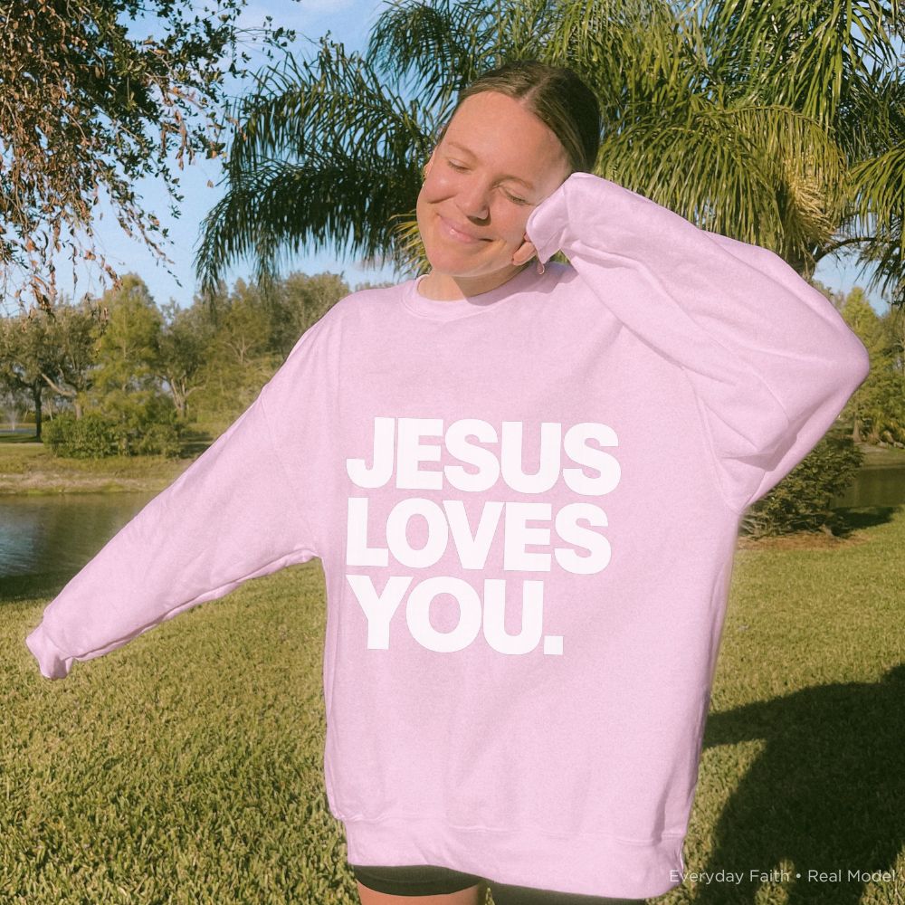 A person outdoors on a sunny lawn wearing a light pink Everyday Faith crewneck sweatshirt with the text 'JESUS LOVES YOU.' printed in bold white capital letters across the front.