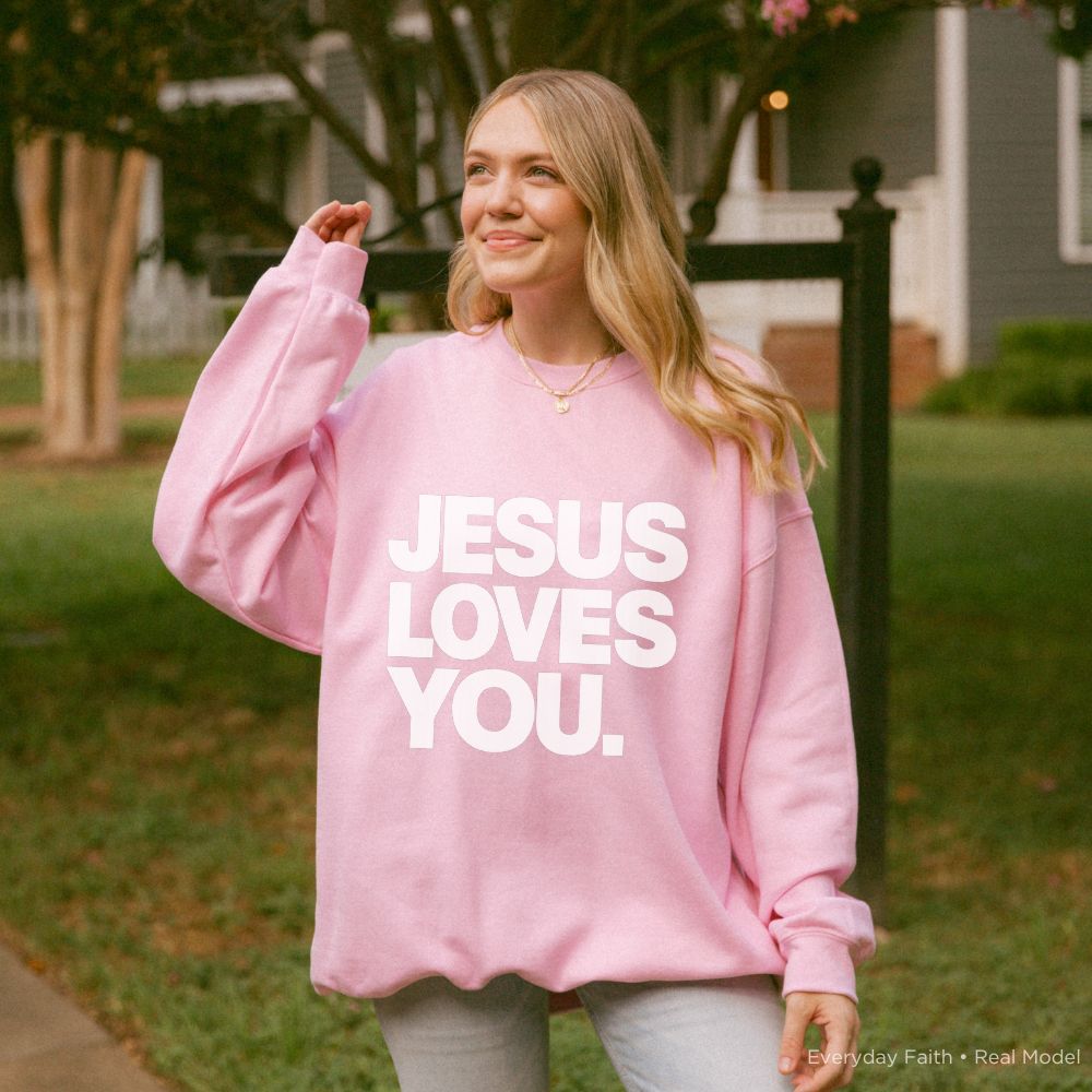 A person wearing a pink Everyday Faith sweatshirt with the text 'JESUS LOVES YOU.' printed in bold white letters, standing outdoors in a grassy yard.