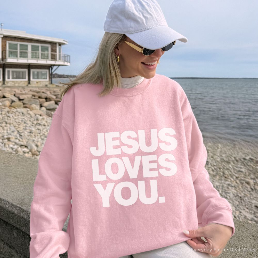 A person wearing a light pink Everyday Faith crewneck sweatshirt with the text 'JESUS LOVES YOU.' printed in bold white capital letters across the front.