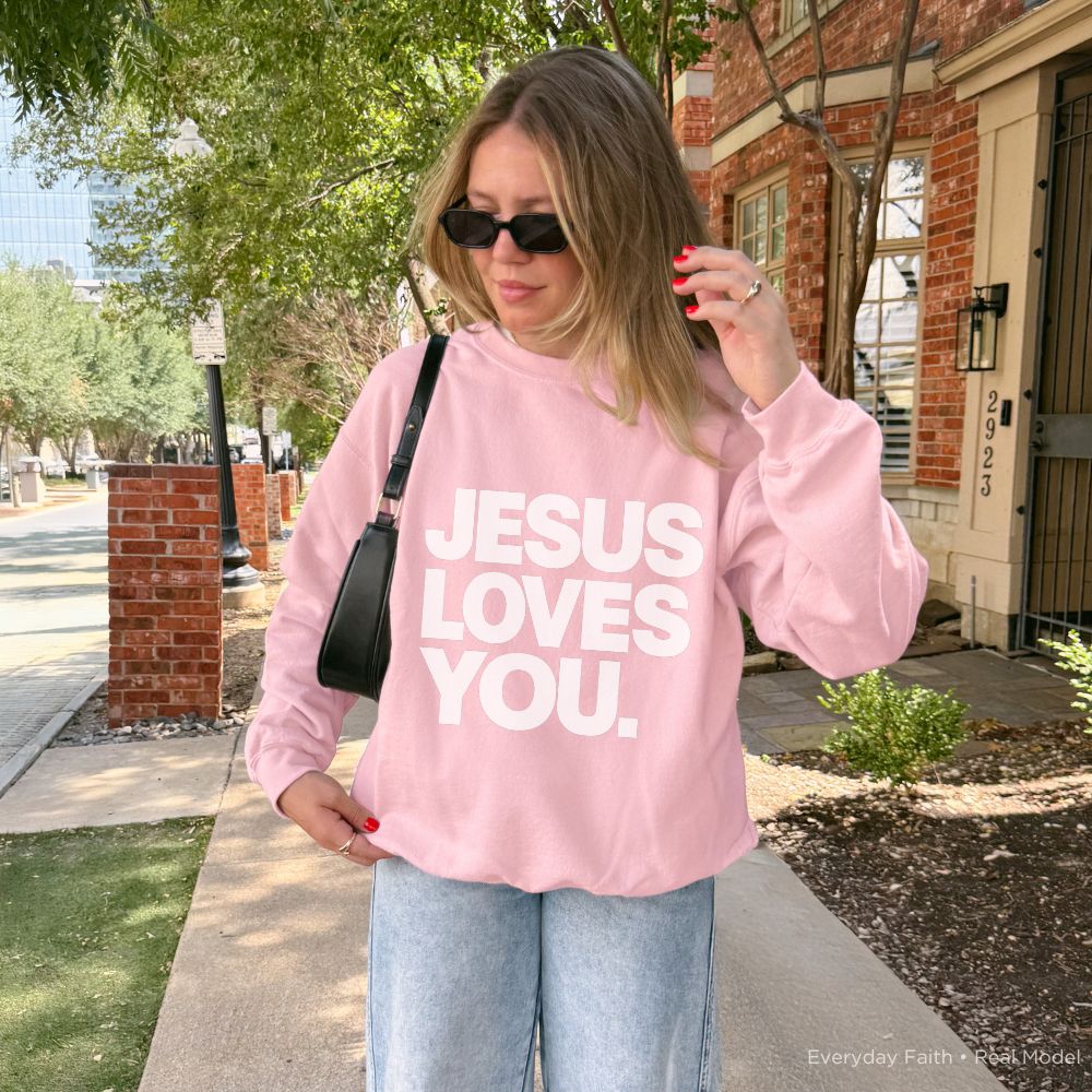 A person wearing a light pink Everyday Faith sweatshirt with the words 'JESUS LOVES YOU.' printed in bold white letters, standing outdoors on a sidewalk in front of a brick building.