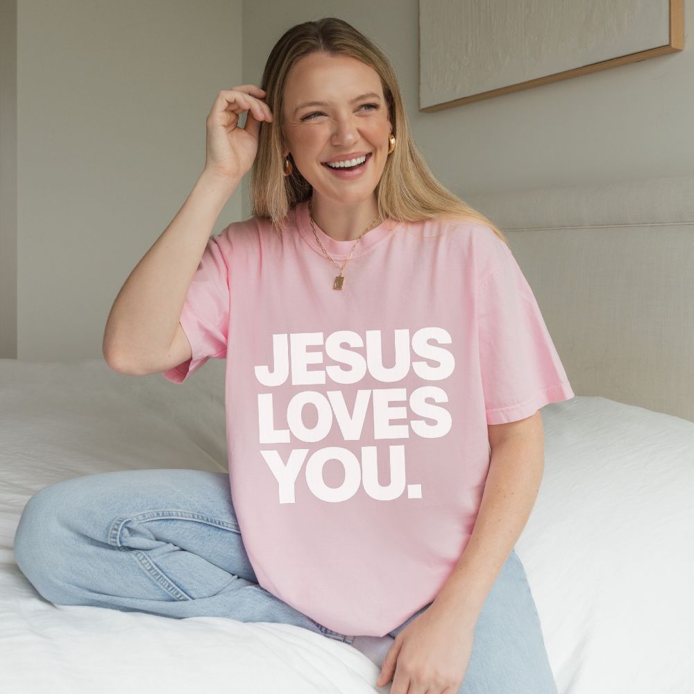 A woman sitting on a bed wearing a light pink Everyday Faith oversized T-shirt with the phrase 'JESUS LOVES YOU.' printed in large white block letters.