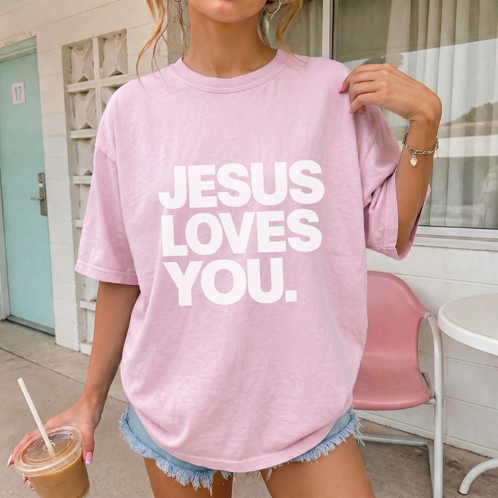 A woman wearing an oversized light pink Everyday Faith t-shirt with the text 'JESUS LOVES YOU.' printed in bold white letters, paired with denim shorts and holding an iced coffee.