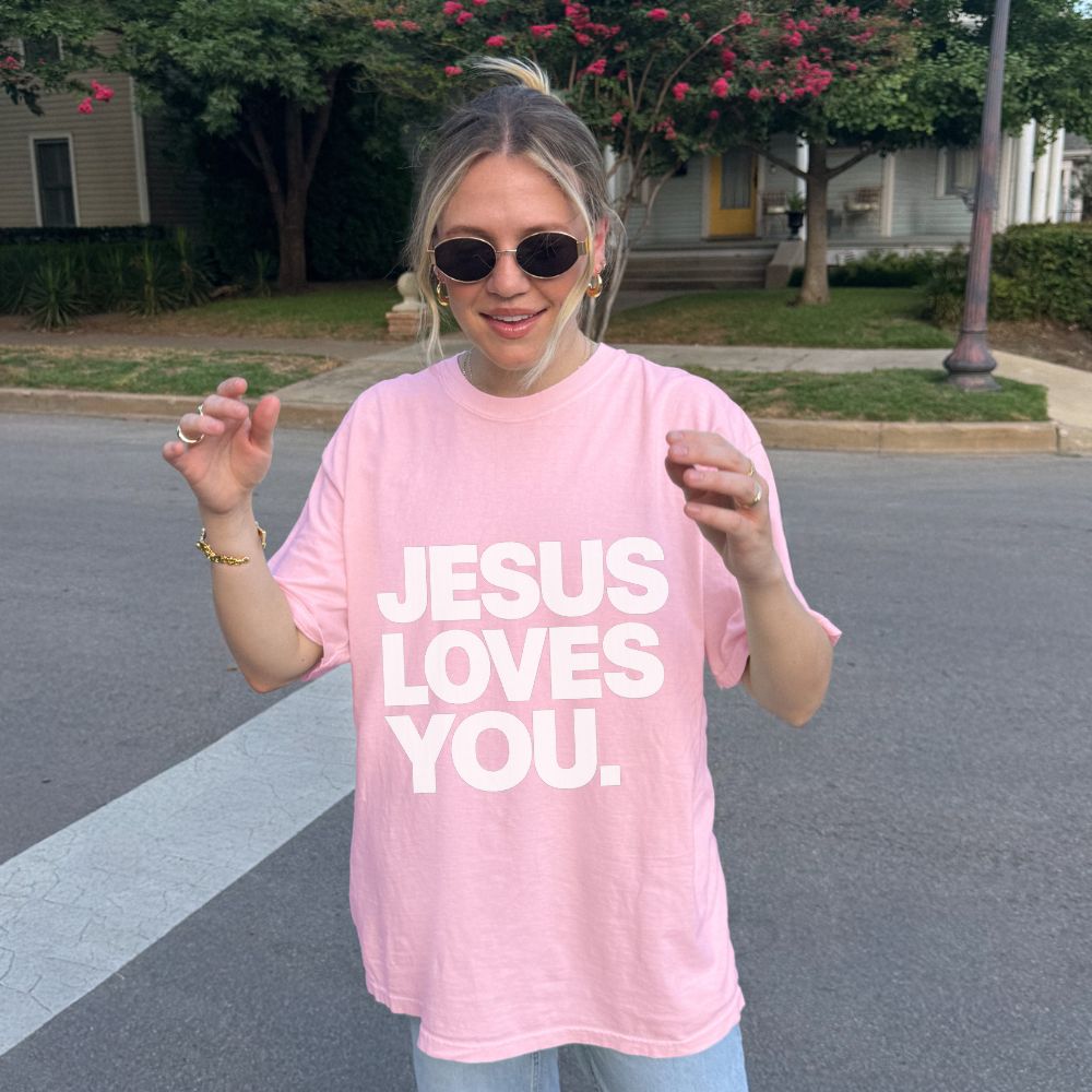 A woman wearing a light pink Everyday Faith oversized t-shirt with the text 'JESUS LOVES YOU.' printed in bold white capital letters, standing outdoors on a paved street with trees and houses in the background.