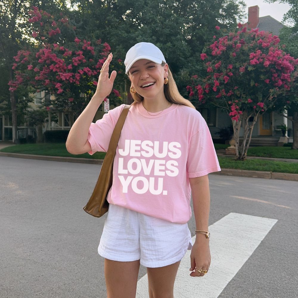 A woman standing on a suburban street crosswalk wearing a light pink Everyday Faith heavyweight t-shirt with the text 'JESUS LOVES YOU.' in bold white letters, white shorts, a white baseball cap, and a brown shoulder bag.