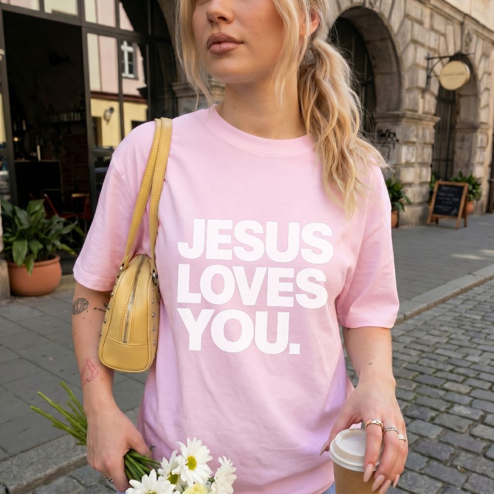 A woman wearing a light pink Everyday Faith Heavyweight t-shirt with the bold white text 'JESUS LOVES YOU.' printed on the front, carrying a small yellow shoulder bag and holding a coffee cup in an outdoor street setting.