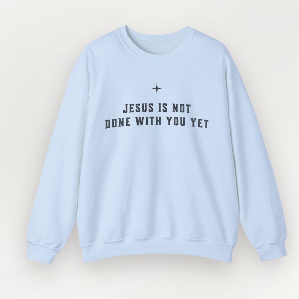 A light blue Everyday Faith crewneck sweatshirt with the text 'JESUS IS NOT DONE WITH YOU YET' printed in black across the chest, displayed on a clean, light beige background.