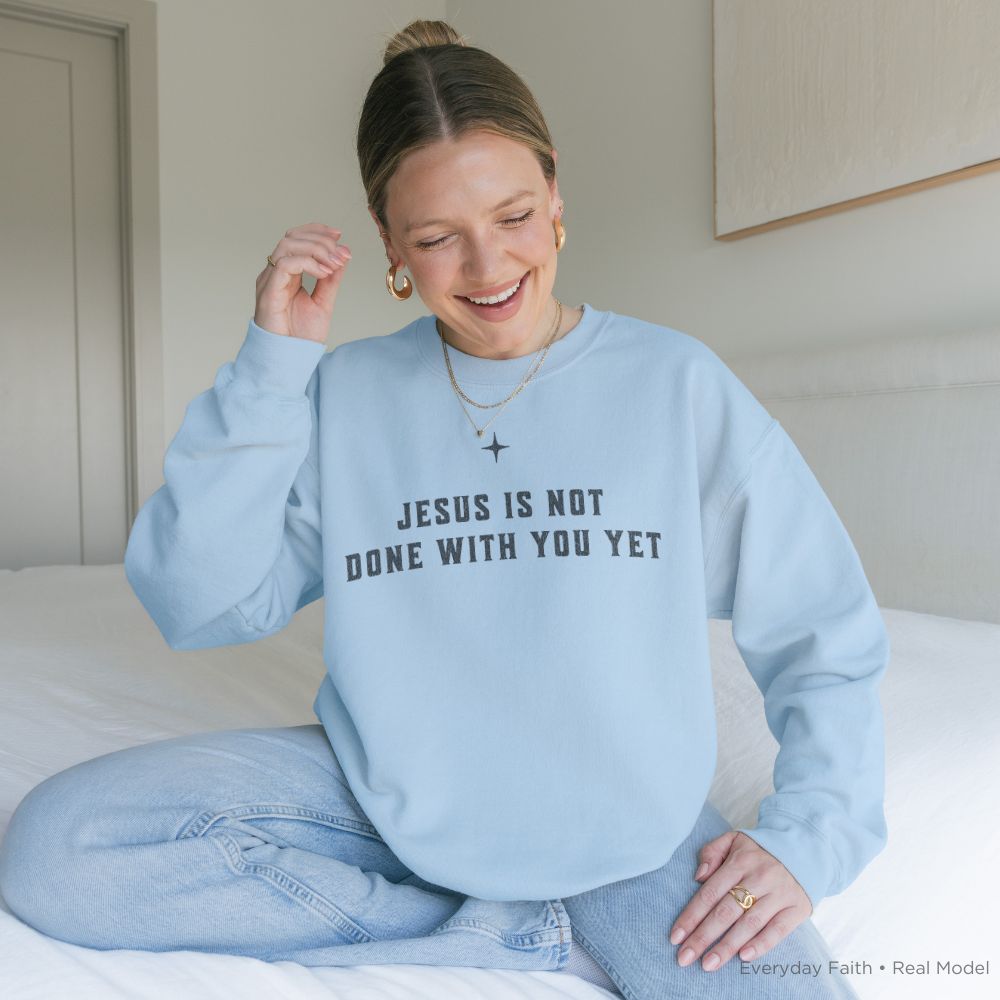 A woman sitting on a bed wearing a light blue Everyday Faith crewneck sweatshirt with the text 'JESUS IS NOT DONE WITH YOU YET' in black capital letters, paired with light-wash jeans.