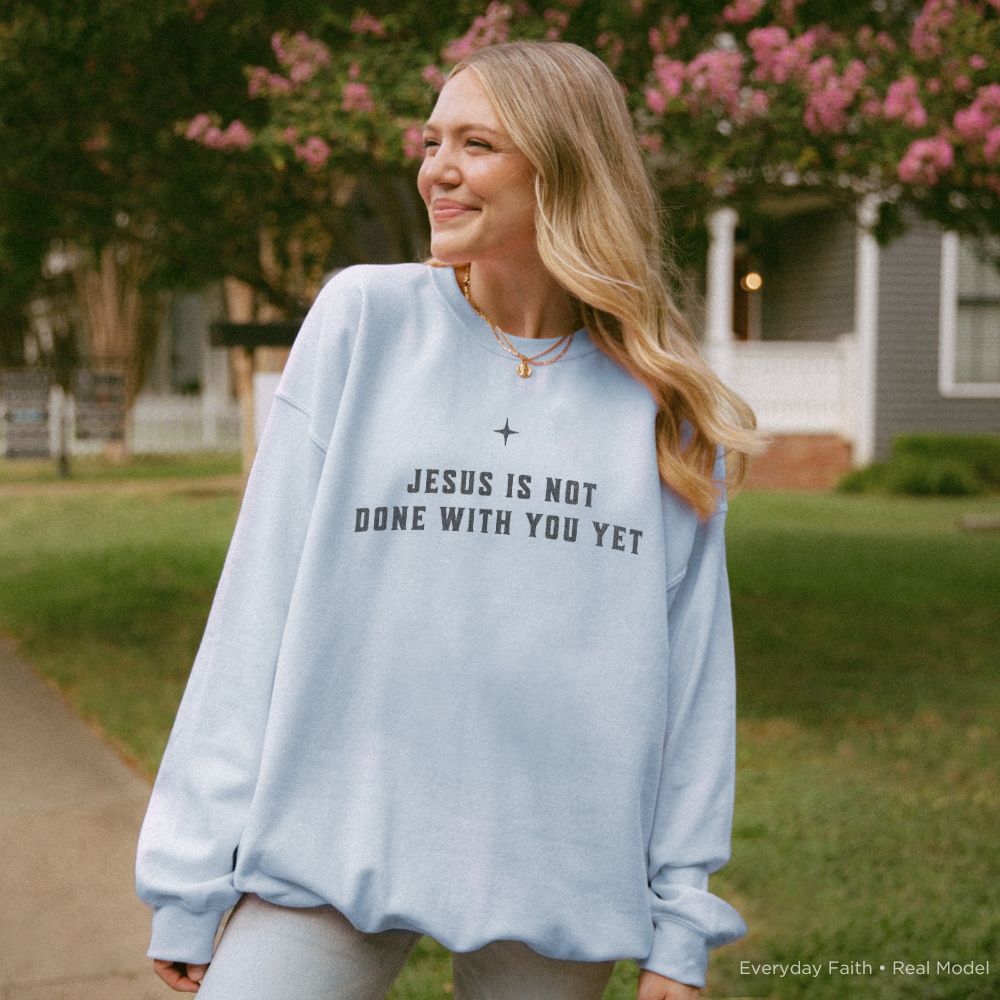 A woman wearing an oversized light blue Everyday Faith sweatshirt with the text 'JESUS IS NOT DONE WITH YOU YET' and a small cross symbol, standing outdoors in a residential neighborhood.