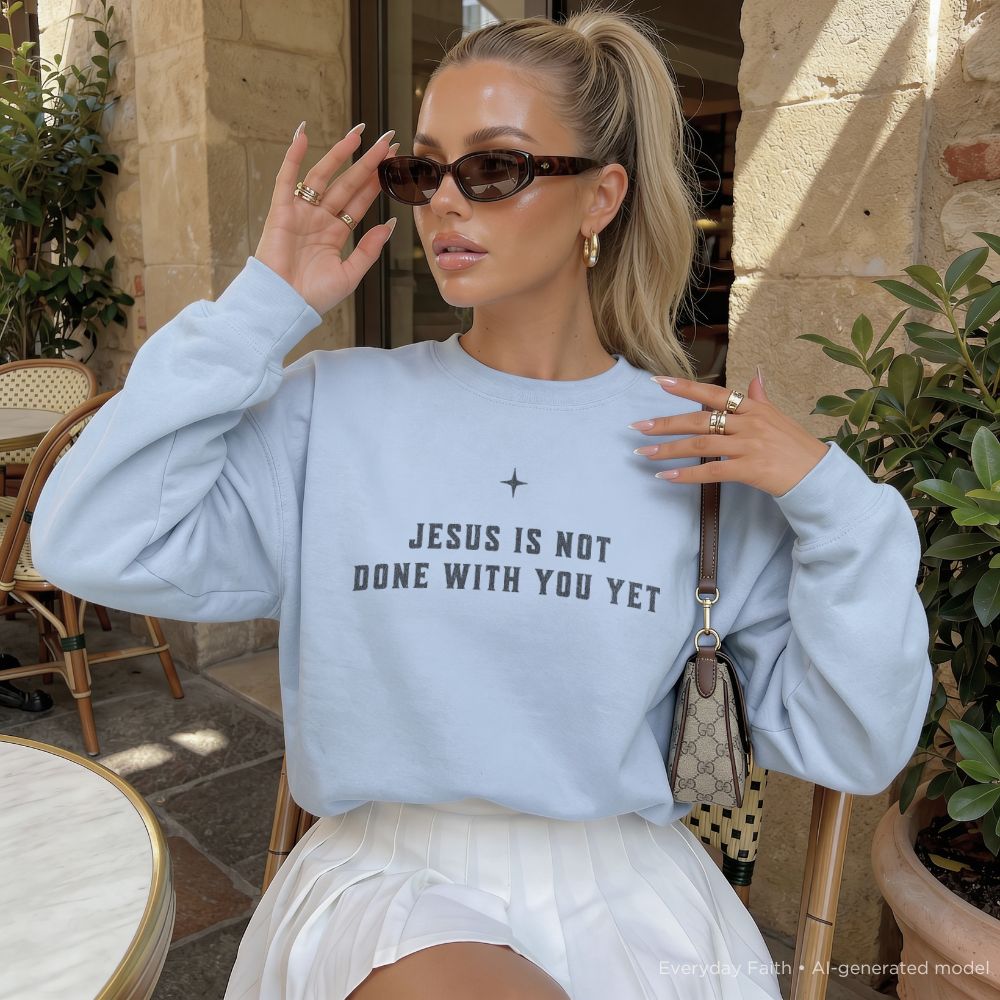 A woman wearing a light blue Everyday Faith crewneck sweatshirt with the text 'JESUS IS NOT DONE WITH YOU YET' in dark gray lettering, paired with a white skirt and sunglasses, sitting in an outdoor cafe setting.