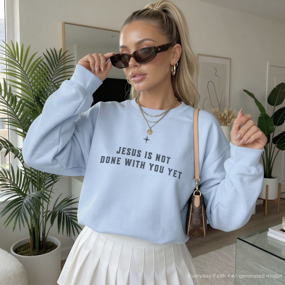 A woman wearing a light blue Everyday Faith crewneck sweatshirt with the text 'JESUS IS NOT DONE WITH YOU YET' in dark gray, styled with a white pleated skirt, sunglasses, and a brown crossbody bag in a bright, modern room with potted plants.