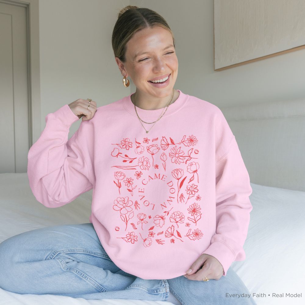 A woman sitting on a bed wearing a light pink Everyday Faith crewneck sweatshirt with a red floral circular graphic and the words 'LOVE CAME DOWN'.