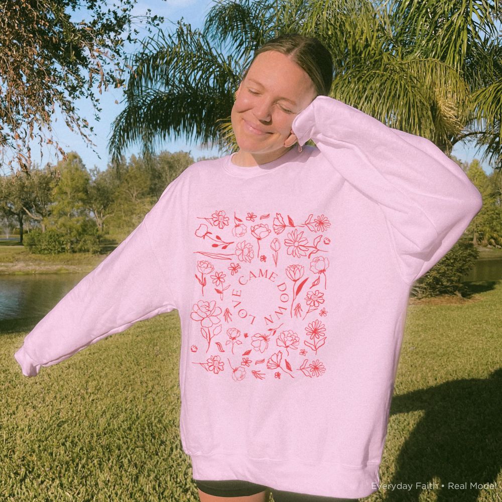 A woman outdoors on a sunny grassy lawn wearing a light pink Everyday Faith crewneck sweatshirt with a red floral graphic and the words 'Love Came down' printed on the front.