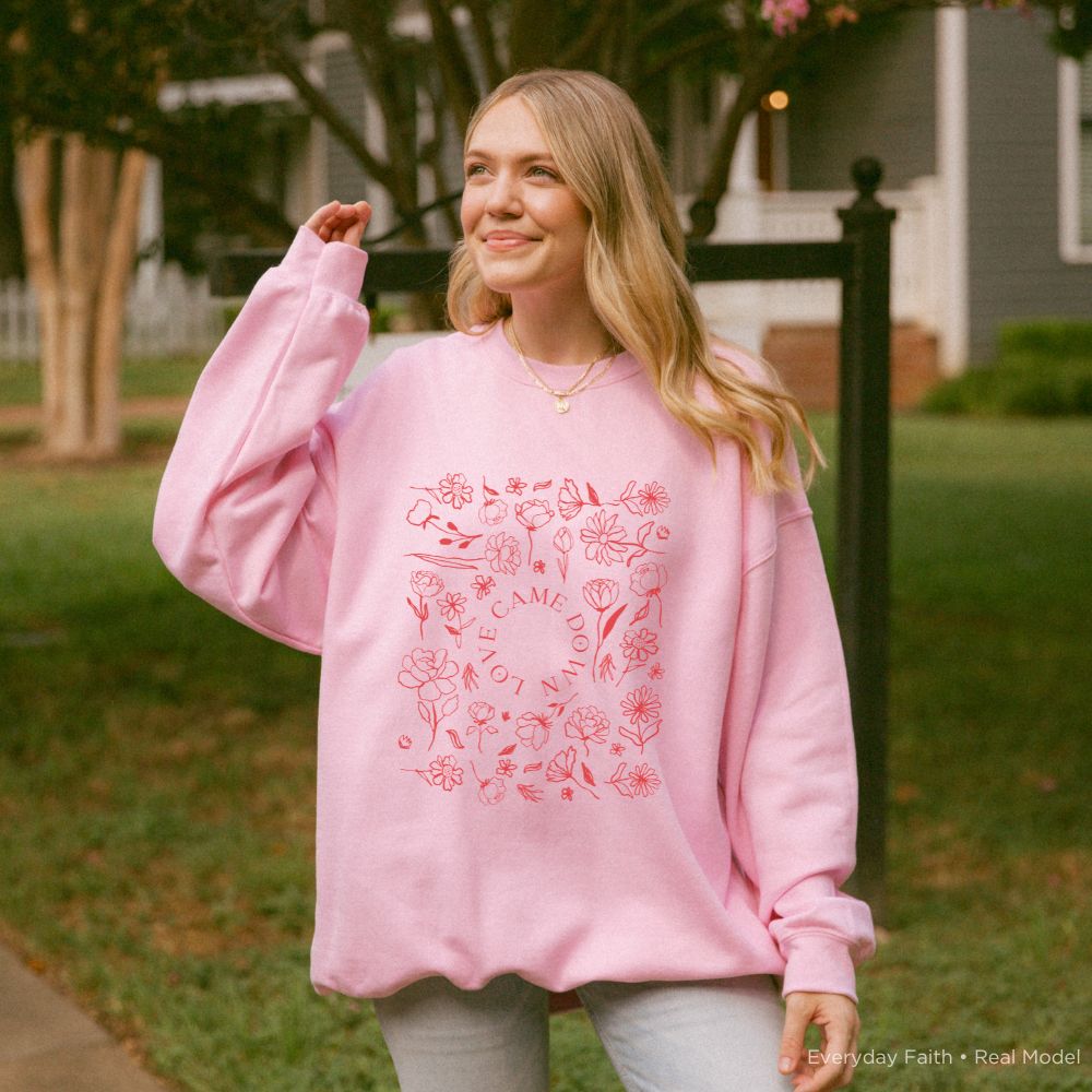 A woman wearing an oversized Everyday Faith light pink sweatshirt with a red floral and 'love came down' graphic print, standing outdoors in a grassy yard.