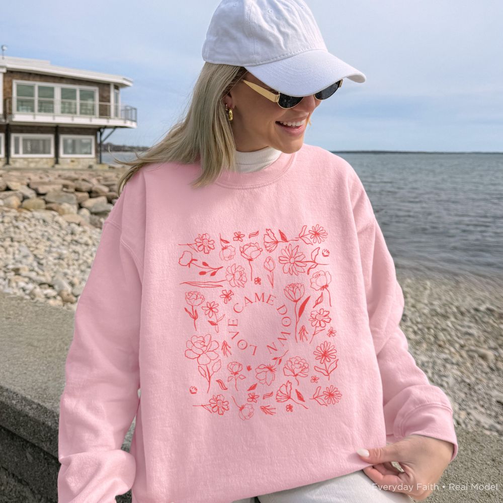 A woman wearing a light pink Everyday Faith crewneck sweatshirt with a red floral graphic and the words 'LOVE CAME DOWN' stands on a rocky shoreline with water and a house in the background.