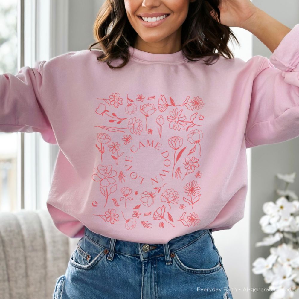 A woman wearing a light pink Everyday Faith crewneck sweatshirt with a red floral and text graphic on the front, paired with blue jeans in a bright indoor setting.