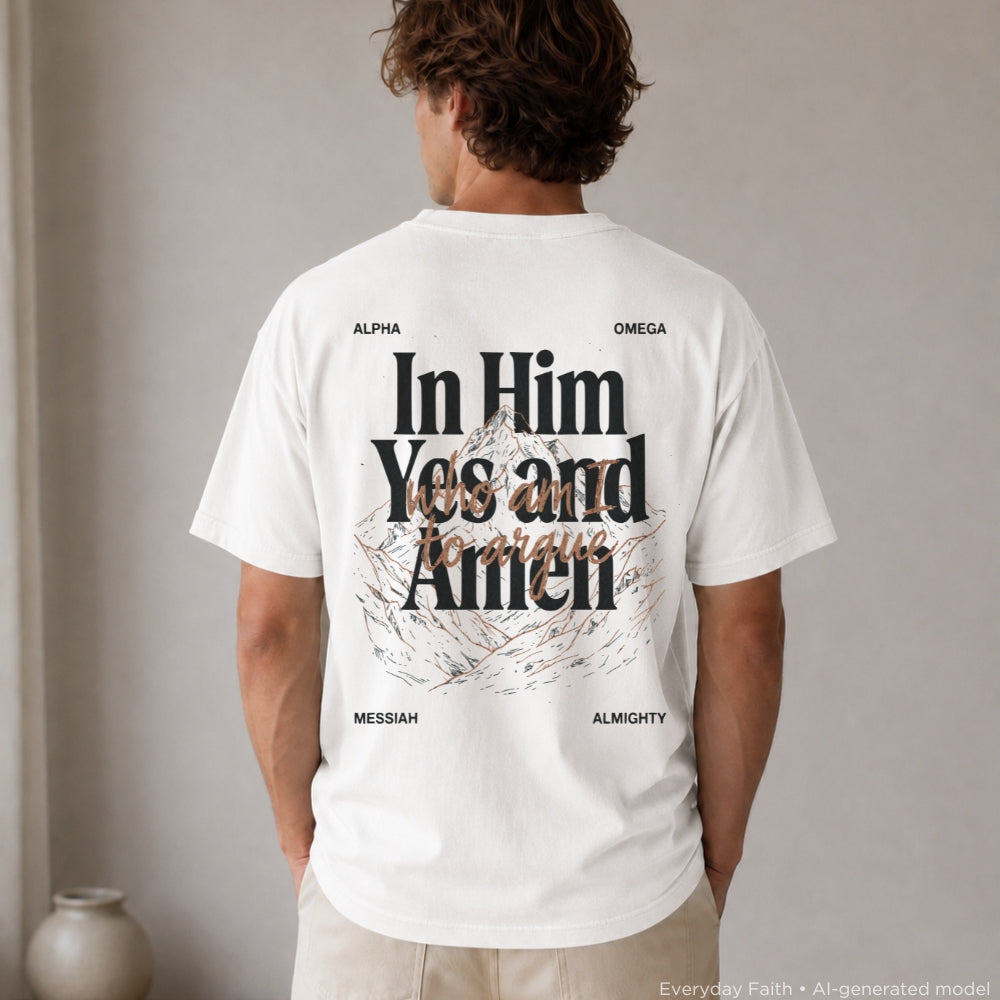 A man viewed from behind wearing a white Everyday Faith graphic T-shirt with large black text reading 'In Him Yes and Amen' and smaller black words around the design, including ALPHA, OMEGA, MESSIAH, and ALMIGHTY. The shirt is shown in a softly lit indoor setting with a neutral wall and a small vase at the lower left.