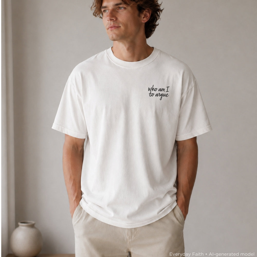 A man wearing a white Everyday Faith crewneck t-shirt with the black cursive text 'who am I to argue' printed on the left chest, paired with beige pants, standing in a minimalist room.
