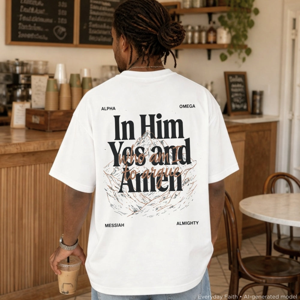 A man viewed from behind wearing a white Everyday Faith graphic t-shirt with a large black and brown print on the back reading 'In Him Yes and Amen' with 'ALPHA' and 'OMEGA' at the top and 'MESSIAH' and 'ALMIGHTY' at the bottom. The setting is a cozy, warm-toned cafe interior.