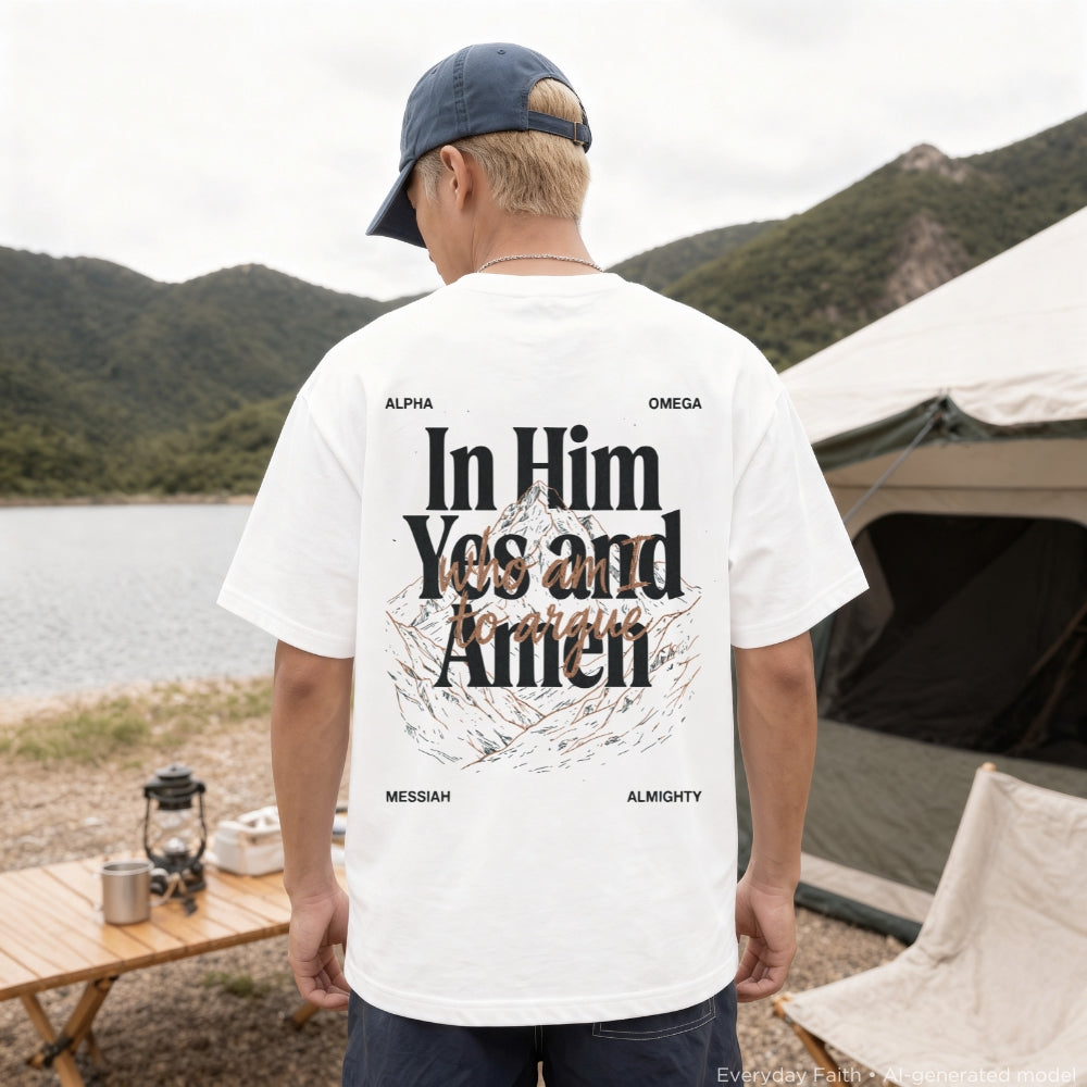 A man stands outdoors at a campsite wearing a white Everyday Faith graphic T-shirt with a large back print reading 'In Him Yes and Amen' and smaller text 'Alpha Omega Messiah Almighty.' Real environment elements like mountains, water, a tent, and a folding table are visible in the background.
