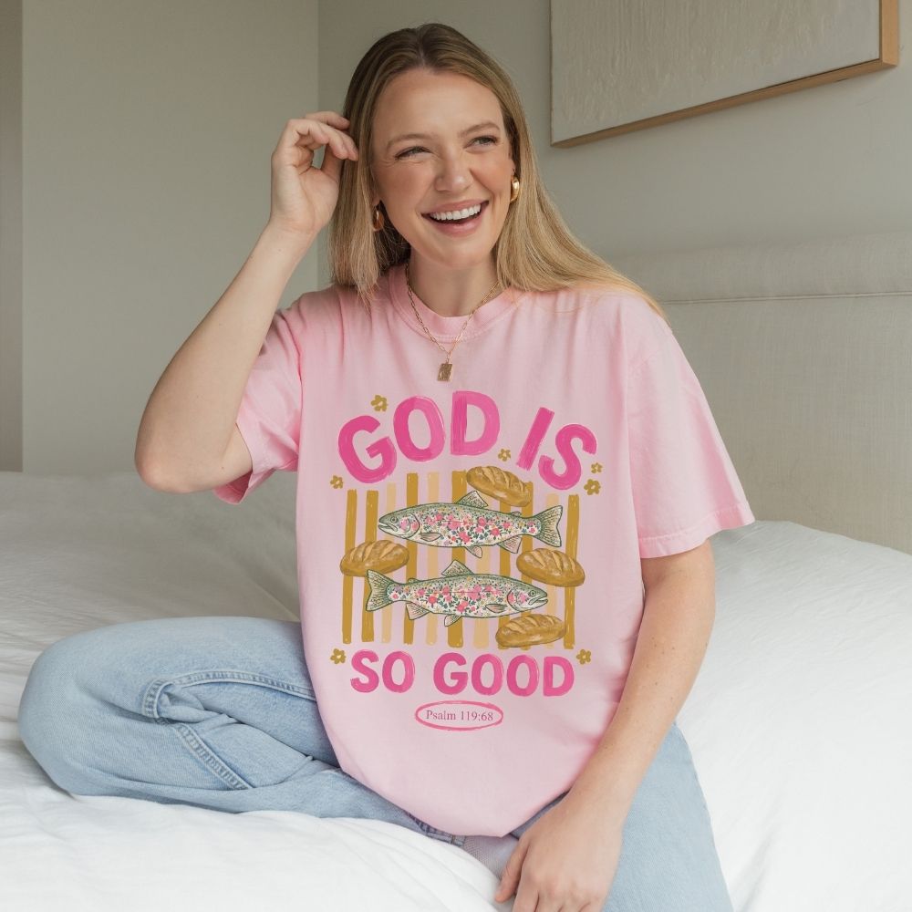 Woman wearing a pink t-shirt with 'God is so good' text sitting on a bed.