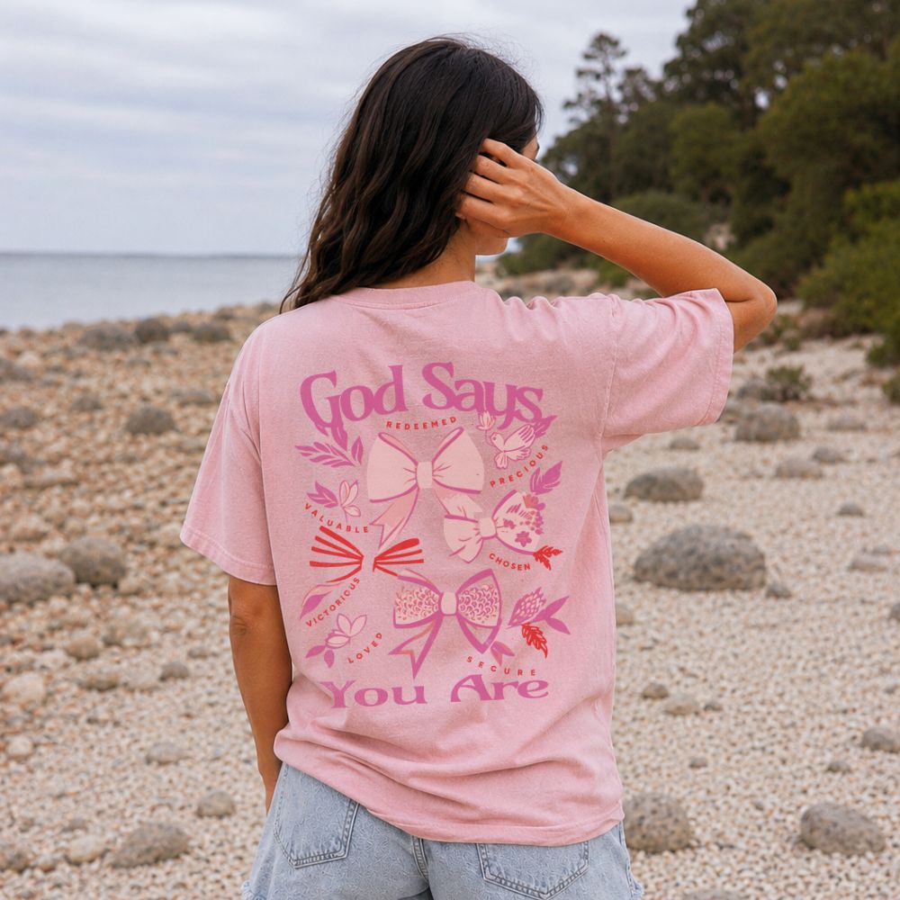 Person wearing a pink Everyday Faith heavyweight t-shirt with text and graphics on a beach. Graphic features pink coquette bows with words ‘God Says You Are’.