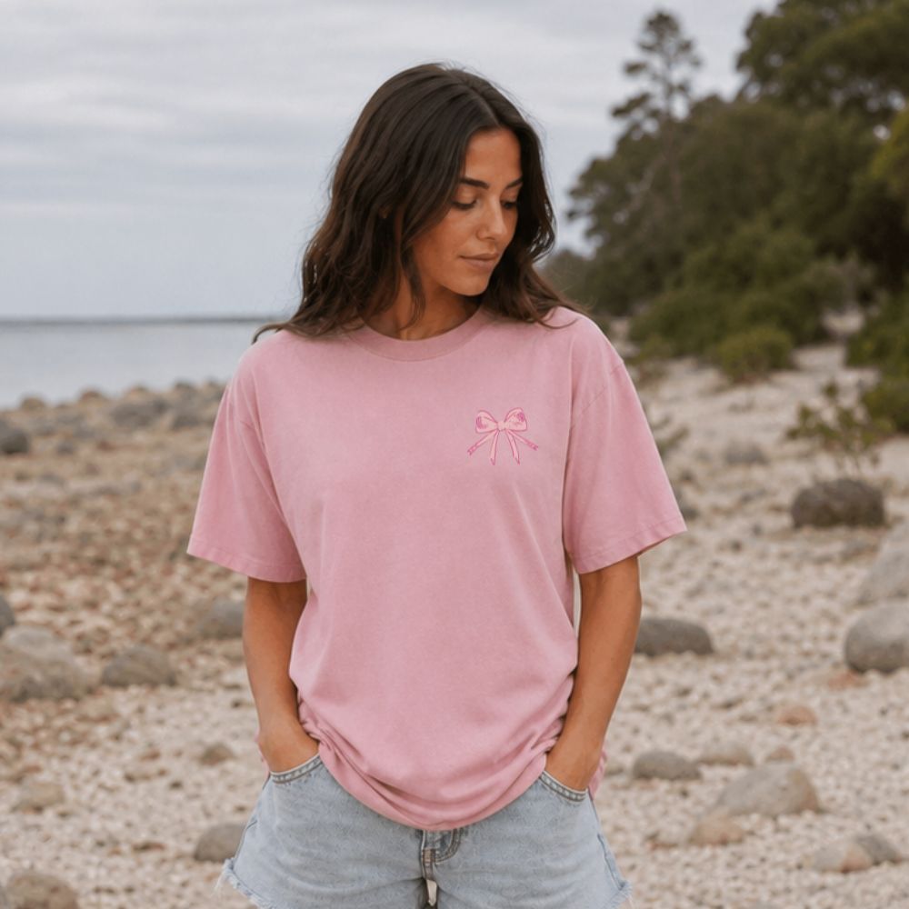 Woman wearing a pink Everyday Faith heavyweight t-shirt with a coquette bow logo on a rocky beach.