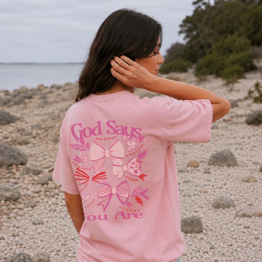 Woman wearing a pink t-shirt with a message on the beach. Graphic features pink coquette bows with words ‘God Says You Are’