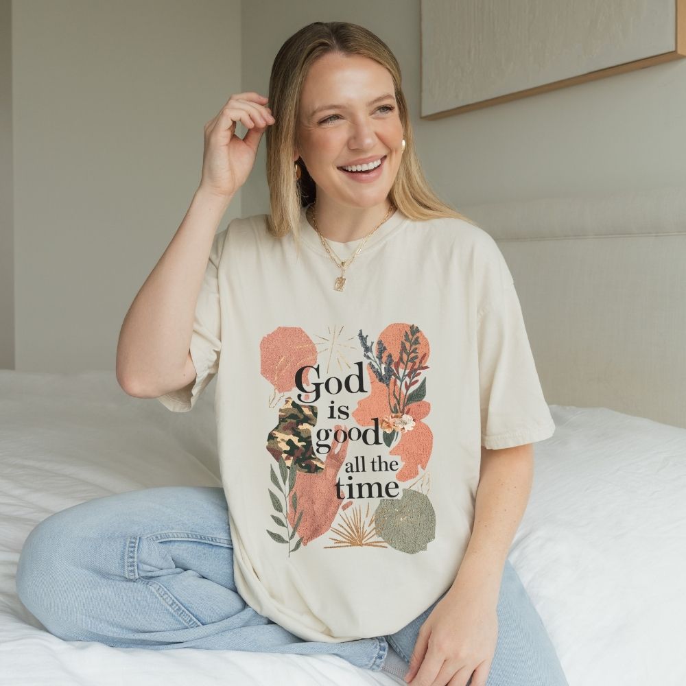 Woman wearing a t-shirt with 'God is good all the time' text and floral design, sitting on a bed.
