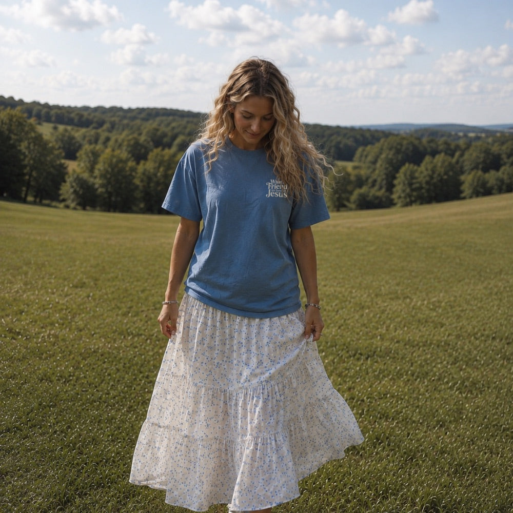 Woman in a blue t-shirt and white floral skirt standing in a grassy field with trees in the background