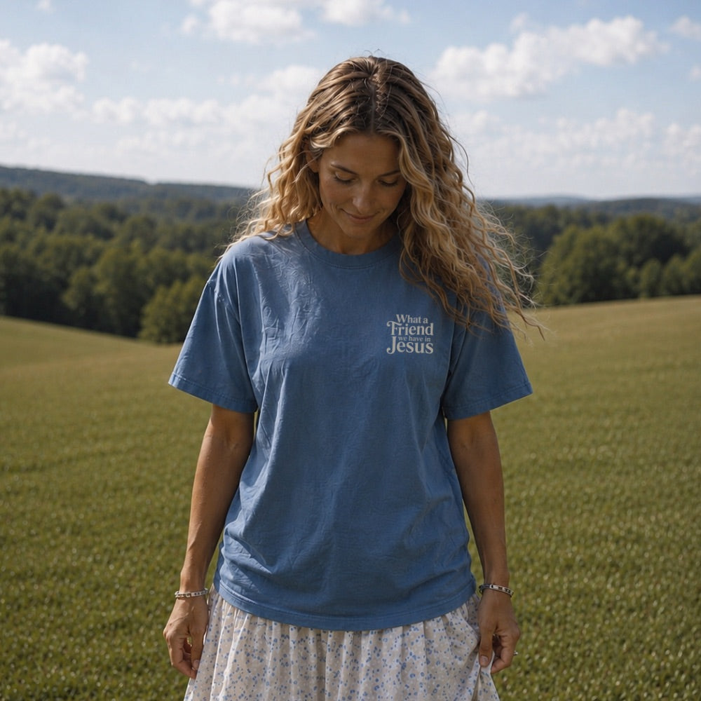 Woman wearing a blue t-shirt with 'What a Friend Jesus' text, standing in a field.