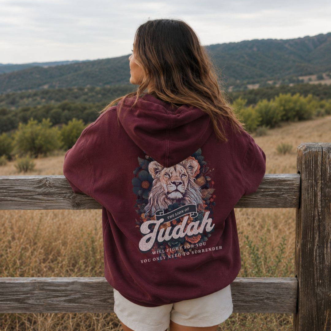 Person wearing a maroon hoodie with a lion design and 'Judah' text, standing by a wooden fence with a scenic background.