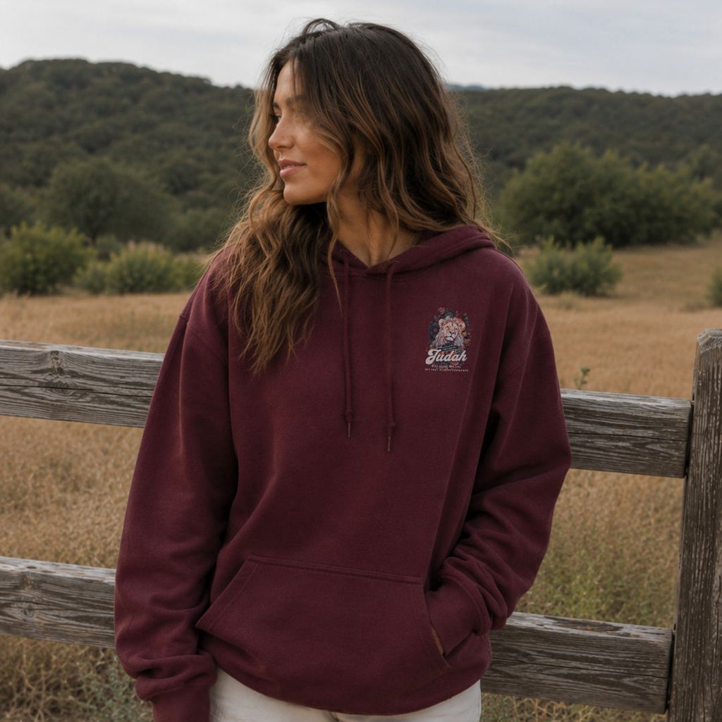 Woman wearing a maroon hoodie with a logo, standing in a field with a wooden fence and trees in the background.