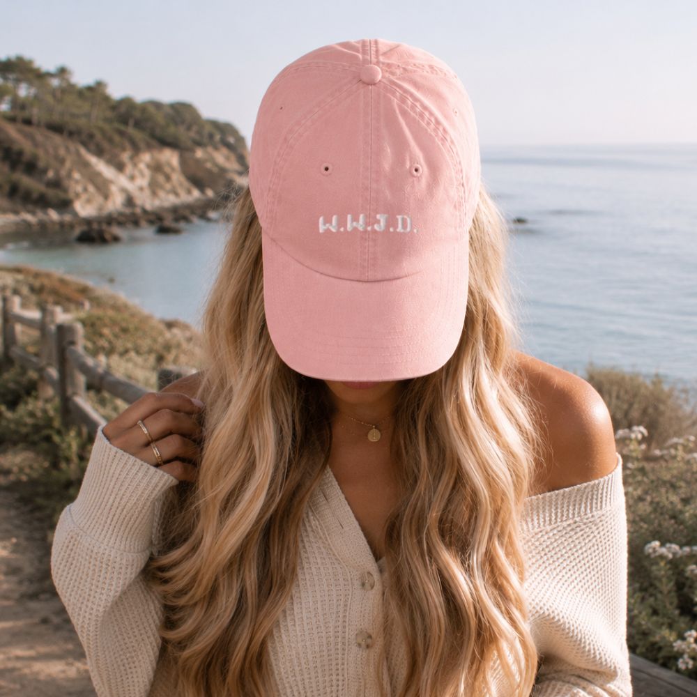 Person wearing a pink cap with 'W.W.I.D.' text, standing by a scenic coastal view.