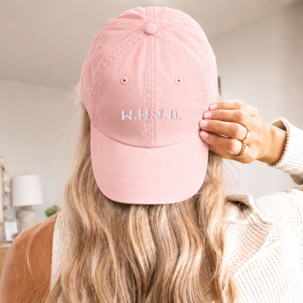 Person wearing a pink cap with 'W.W.J.D.' text, holding it over their head.