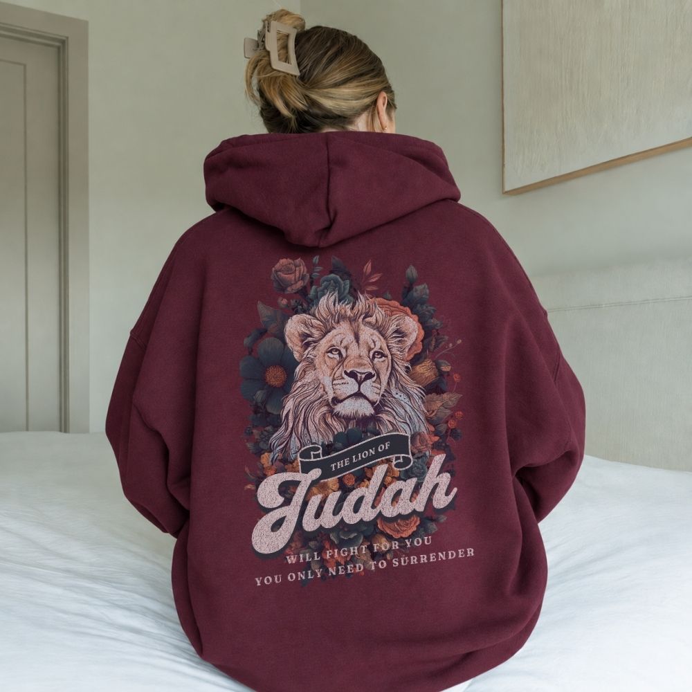 Person wearing a maroon hoodie with a lion graphic and text on the back, sitting on a bed.