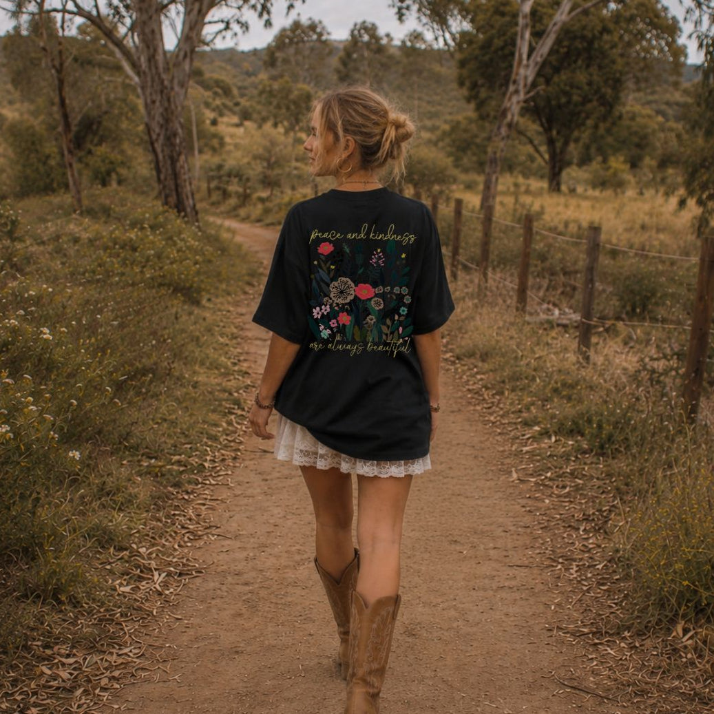 Woman walking on a dirt path in a natural setting wearing a black t-shirt with floral designs and text.