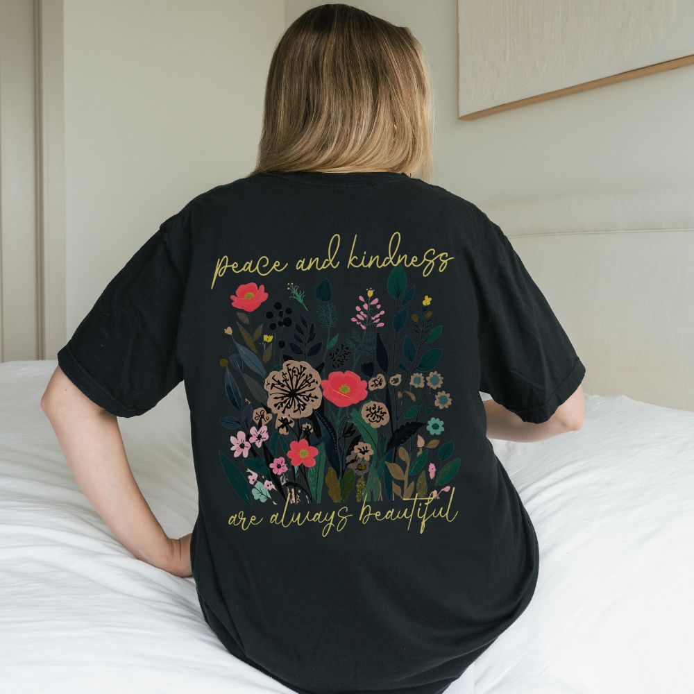 Person wearing a black t-shirt with floral design and text on the back, sitting on a bed.
