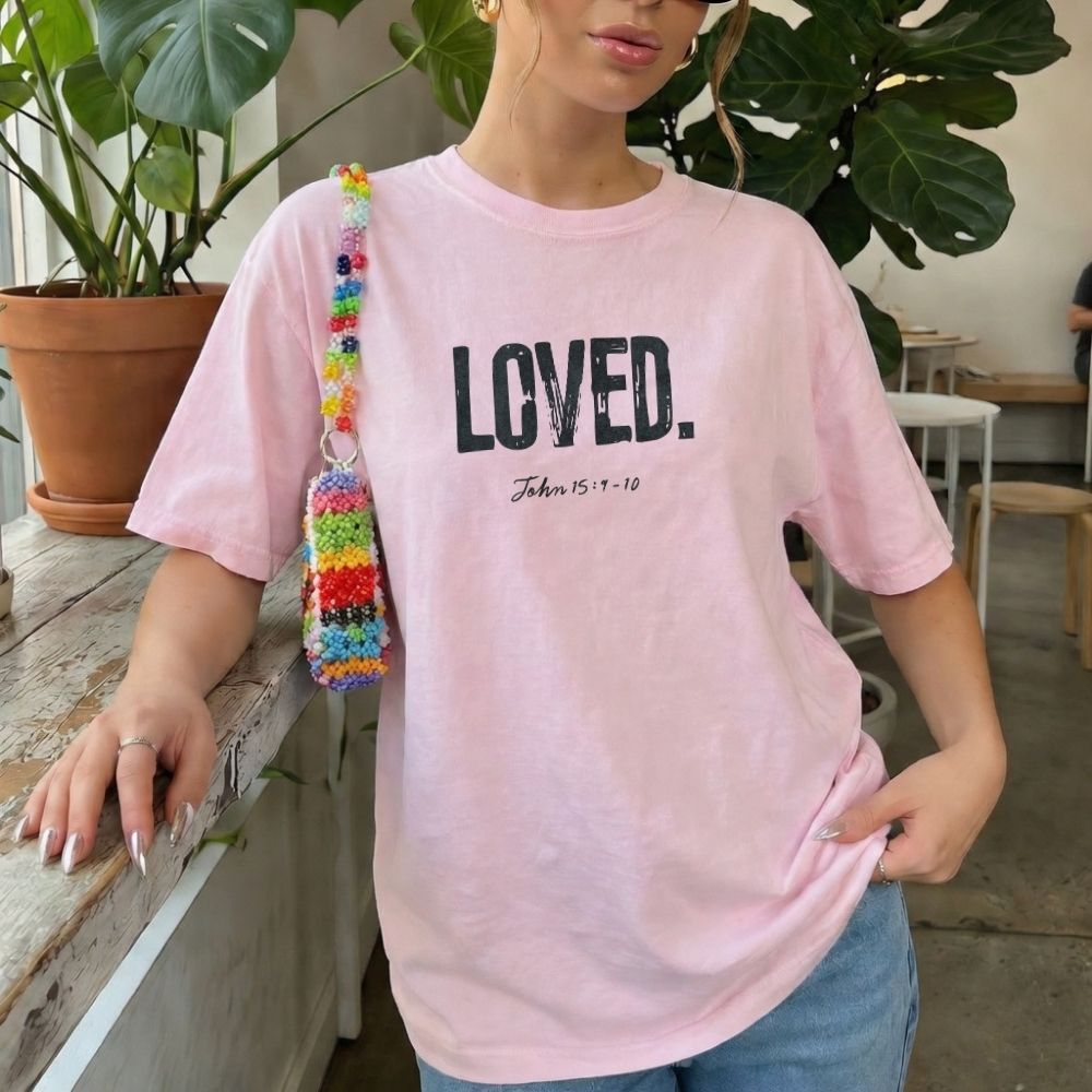 Person wearing a pink t-shirt with 'LOVED' text, standing indoors with plants and furniture in the background.