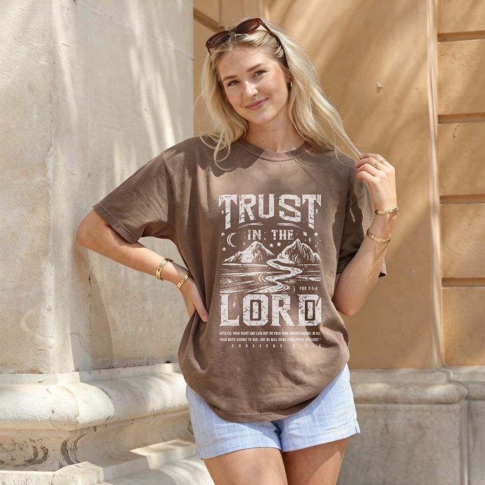 Woman wearing a brown t-shirt with 'Trust in the Lord' text and graphic, standing outdoors.