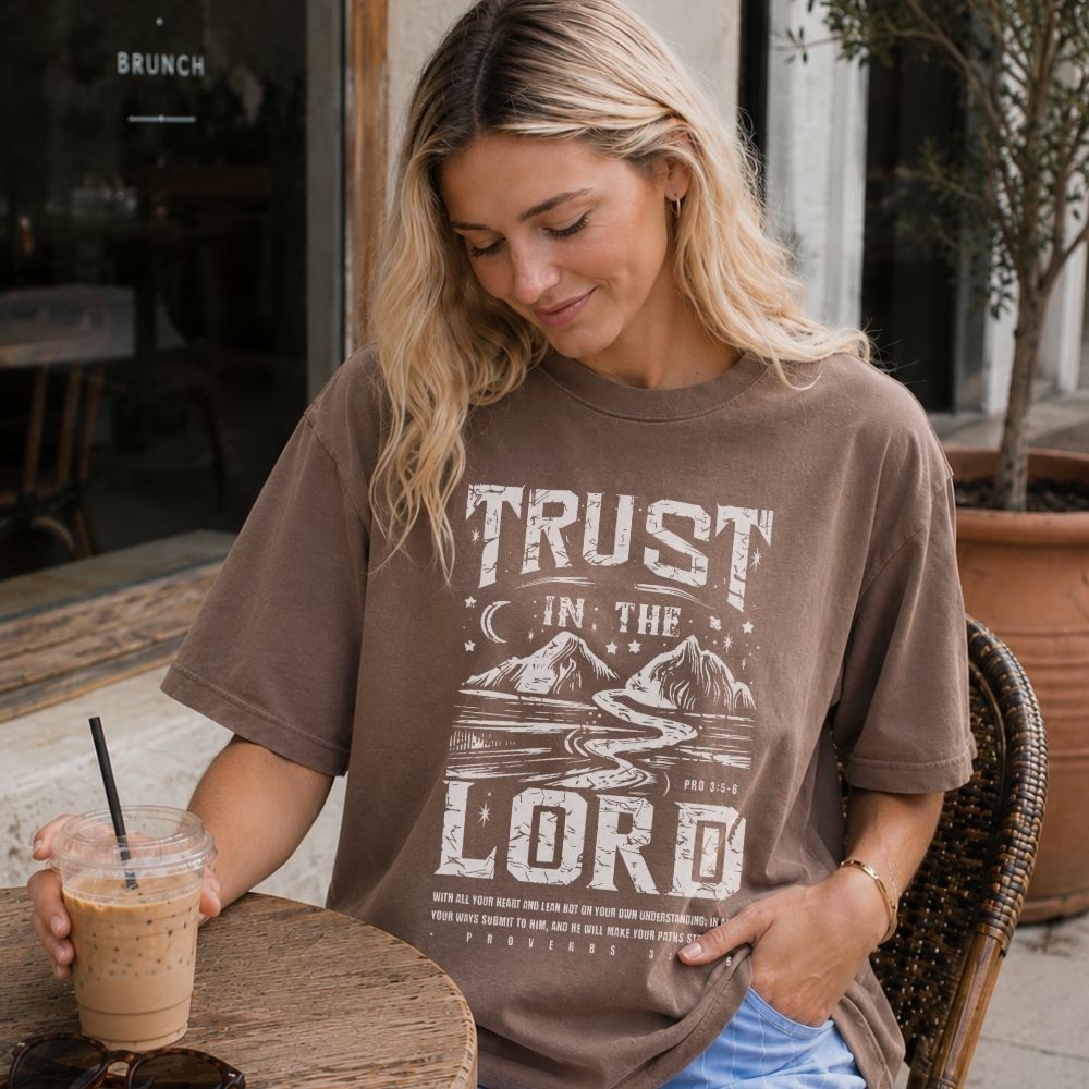 Woman wearing a 'Trust in the Lord' t-shirt, sitting outdoors with an iced coffee.