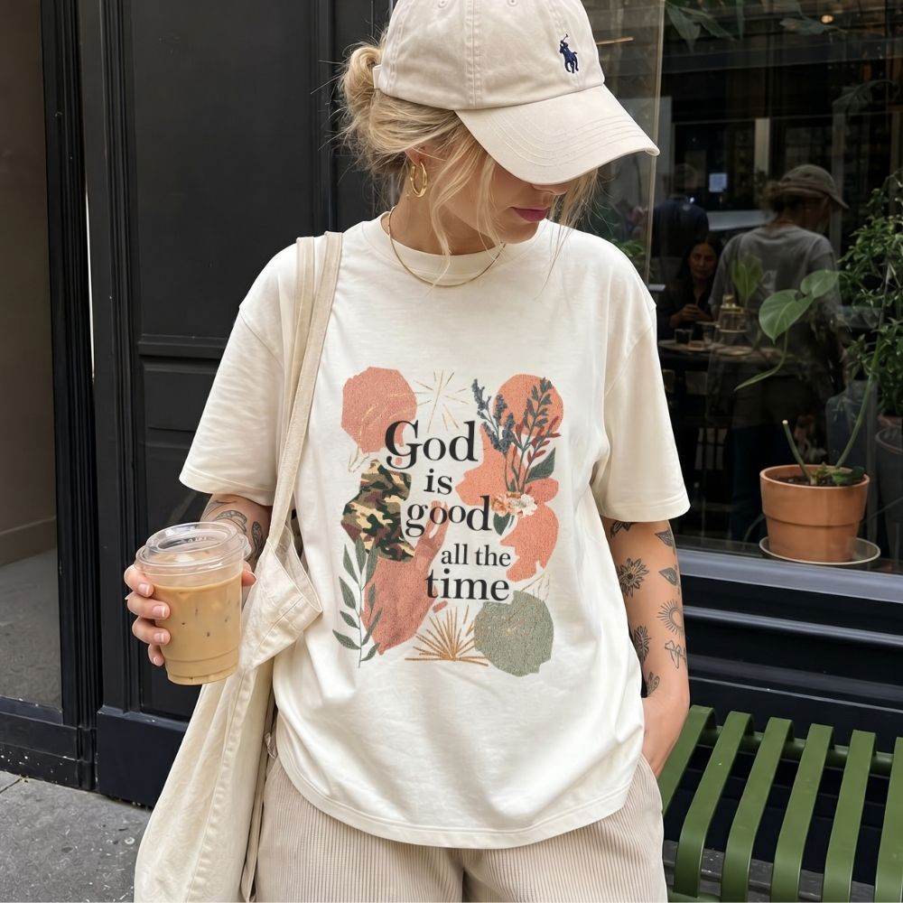Person wearing a white t-shirt with 'God is good all the time' print, holding a drink, standing on a street.