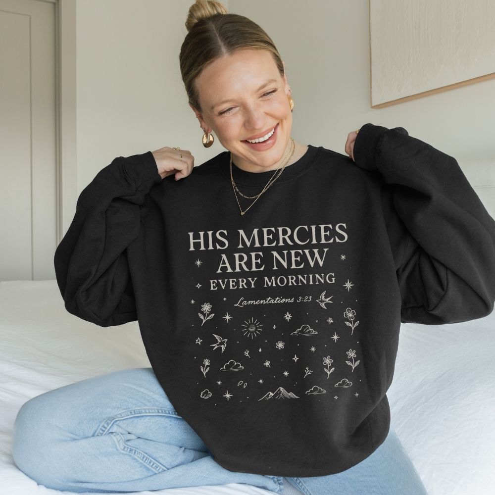 Woman wearing a black sweatshirt with text and graphics, sitting on a bed.