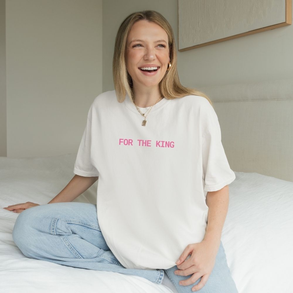 Woman wearing a white t-shirt with 'FOR THE KING' text in pink, sitting on a bed.