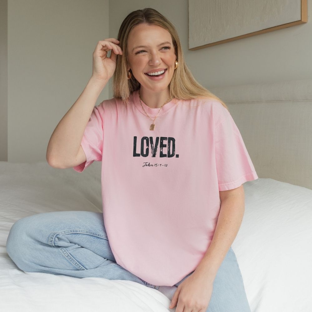 Woman wearing a pink t-shirt with 'LOVED' text, sitting on a bed.